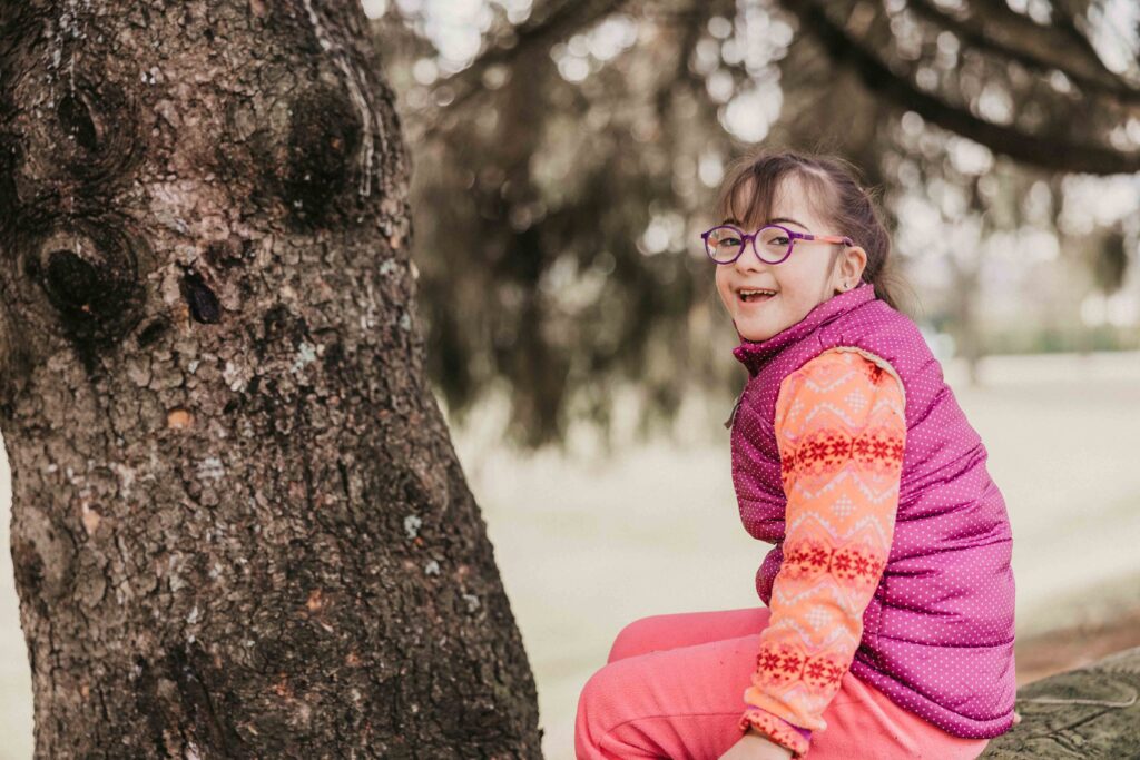 Rebecca in a pink puffy vest sits on a tree trunk smiling over her shoulder at the camera. A green space is visible behind her.