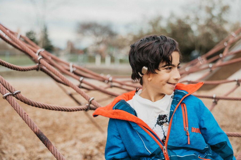 Emilio sits on playground set, smiling at something off camera to the right. He's wearing a blue and orange coat and a t-shirt with a soccer ball on it, and showing off his hearing aid.