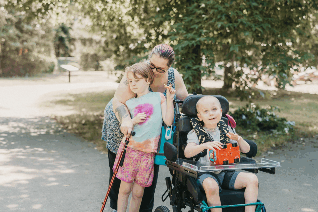 Ender holds a toy truck and smiles. Behind his wheelchair, his mom Sonja leans in close, one arm wrapped around his twin sister, Ellora.