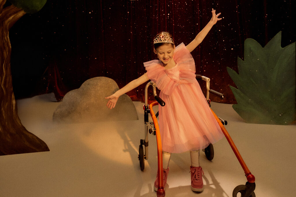Young girl in princess dress and crown dances in front of a stage backdrop. Her walker stands behind her.