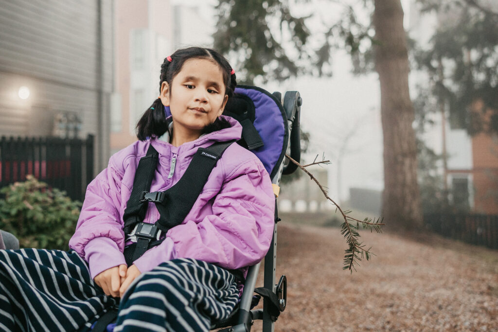 Child sits in a wheelchair on misty path, looking straight at the camera. She's wearing two pony tails and a pink coat.