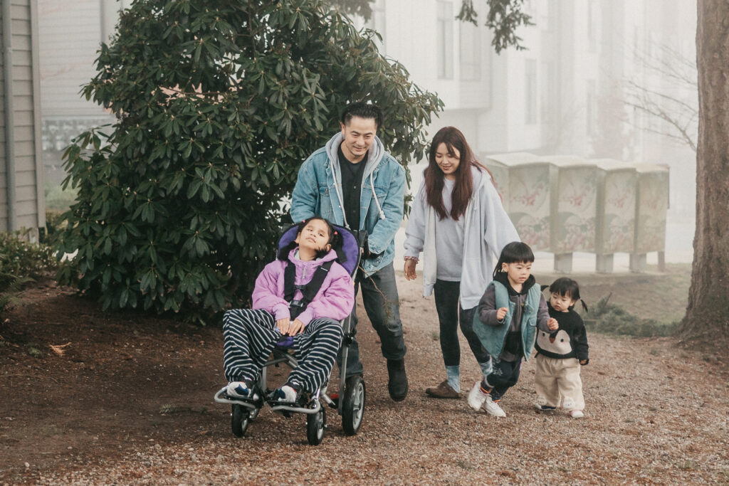 A mom and dad push their daughter in an adaptive stroller. Their two younger kids run beside them. Mist obscures the background.