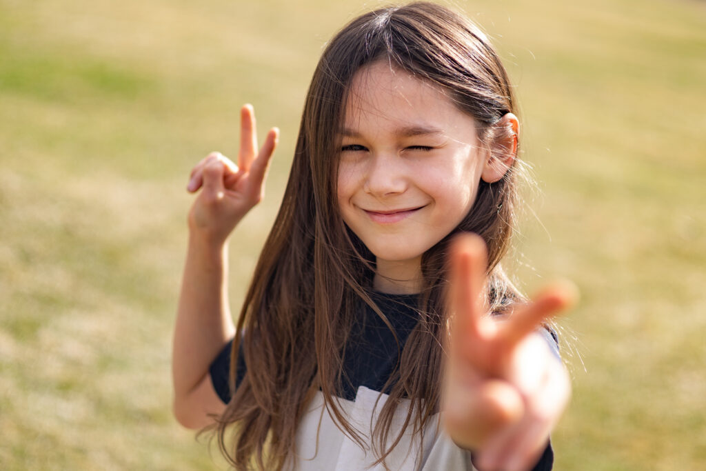 A young boy smiles and squints at the camera, making peace signs with each hand.