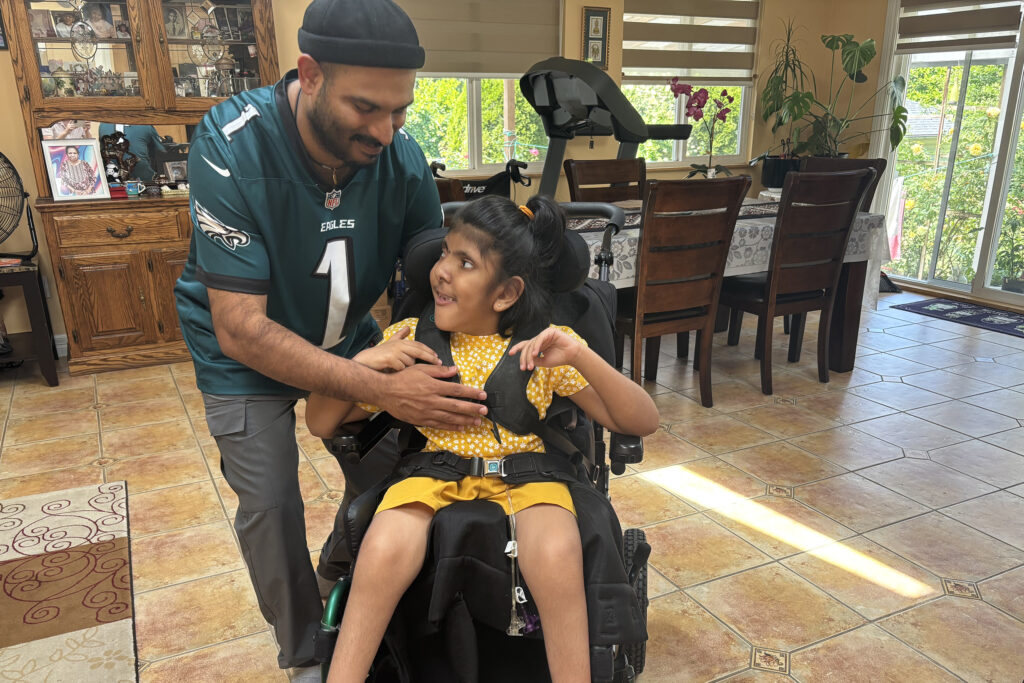 Jaanvi smiles from her wheelchair, head turned toward her dad Sheeru who looks down at her.