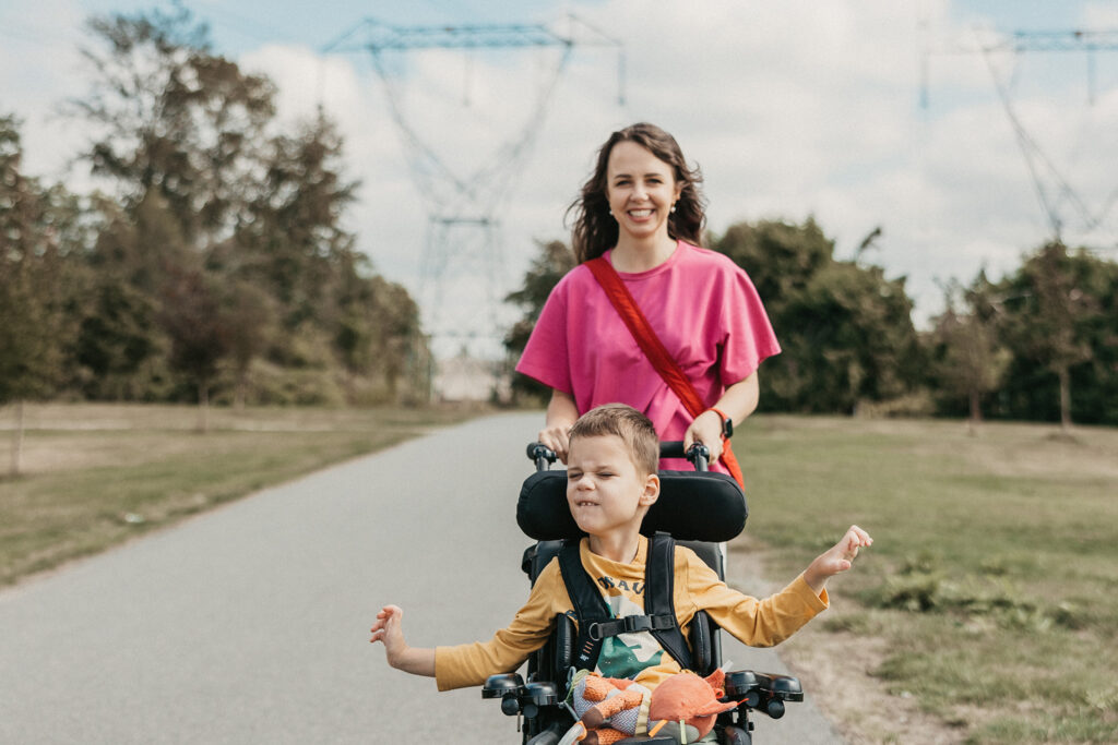 A young boy with cerebral palsy spreads his arms wide and faces the sun. His mom pushes his wheelchair.
