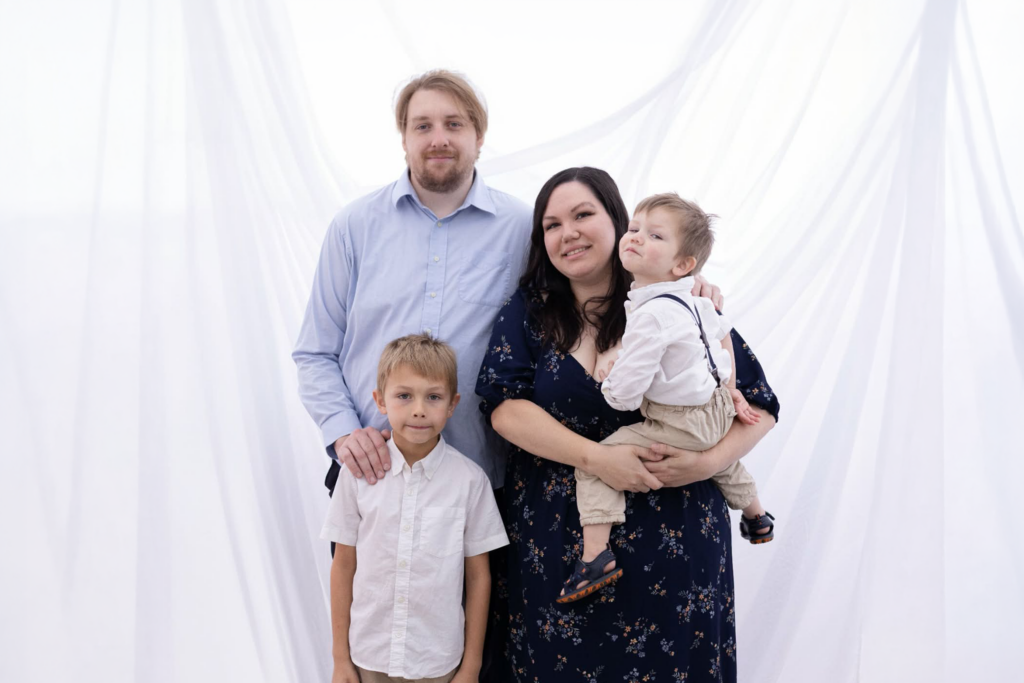 A family with parents and two young boys poses in front of a light background.