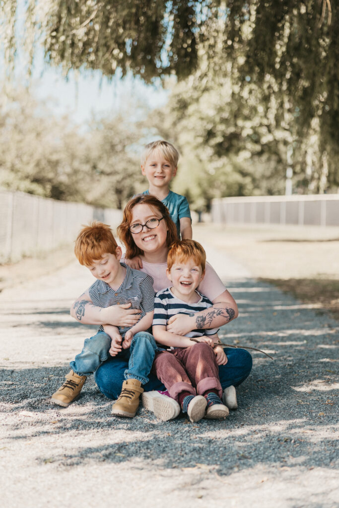 A mother hugging her 3 sons on a sunny day with trees.