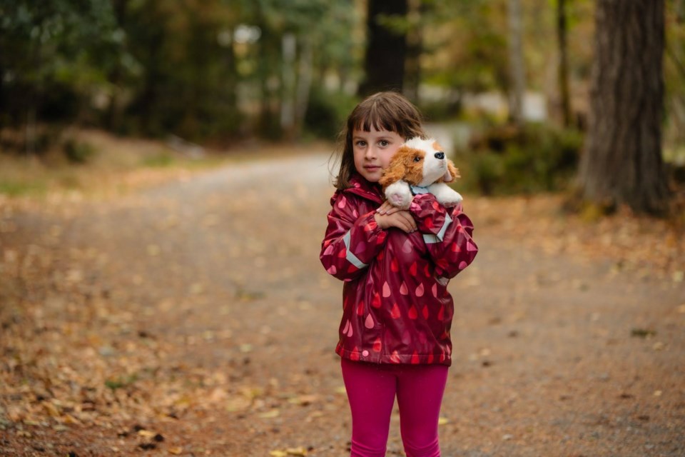A young girl with light brown hair and bangs is wearing a rain jacket and leggings. She is standing on a trail surrounded by trees and holding a small dog stuffed animal.